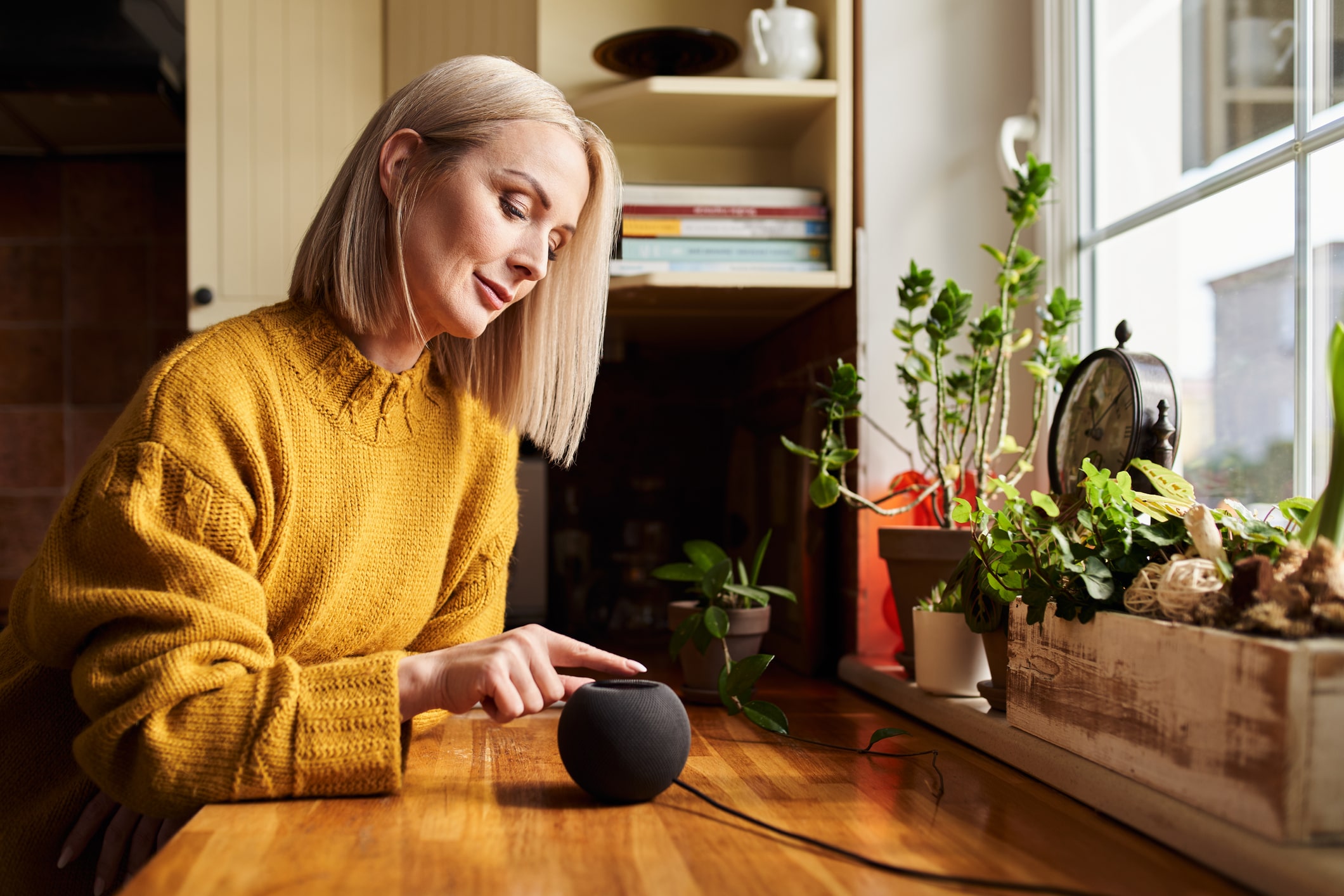 The image shows an elderly woman with gray hair and a yellow sweater leaning against a table and interacting with a voice assistant, an Apple HomePod mini.